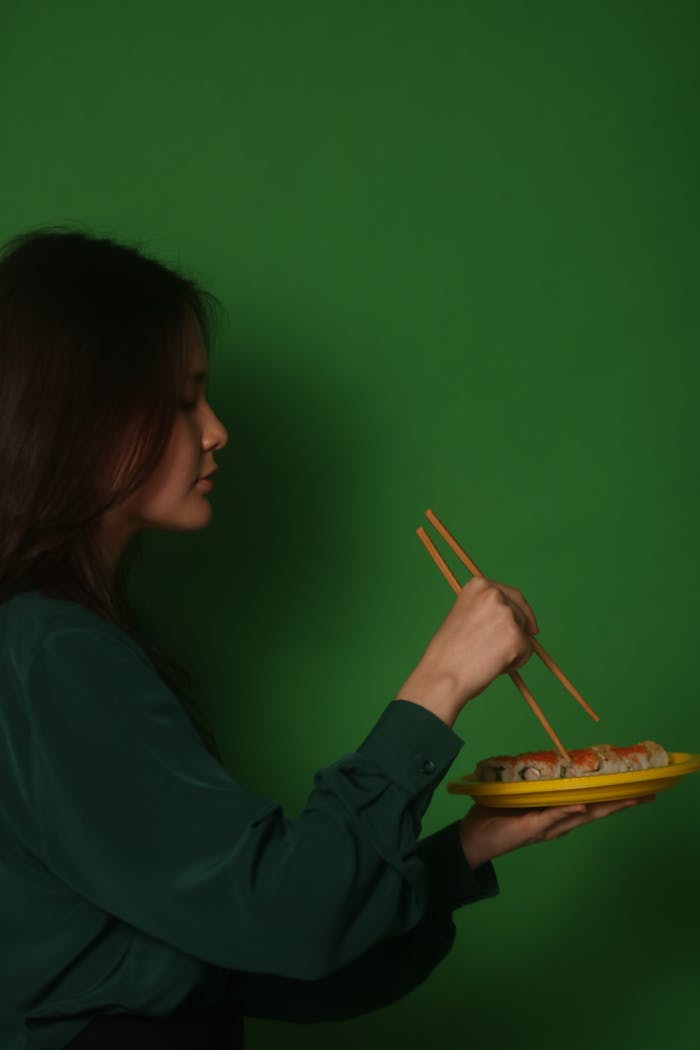 A woman with chopsticks eating sushi rolls against a vibrant green backdrop, showcasing Japanese cuisine.